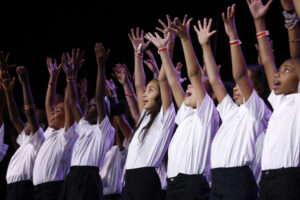 Elementary students singing on stage with their arms raised wearing white shirts
