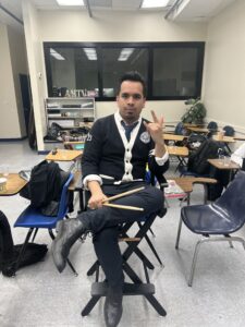 music teacher sitting with drumsticks and giving "I Love You" ASL sign language