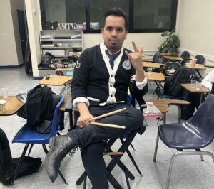 music teacher sitting with drumsticks and giving "I Love You" ASL sign language