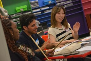 Music teachers seated together smiling and playing drumsticks